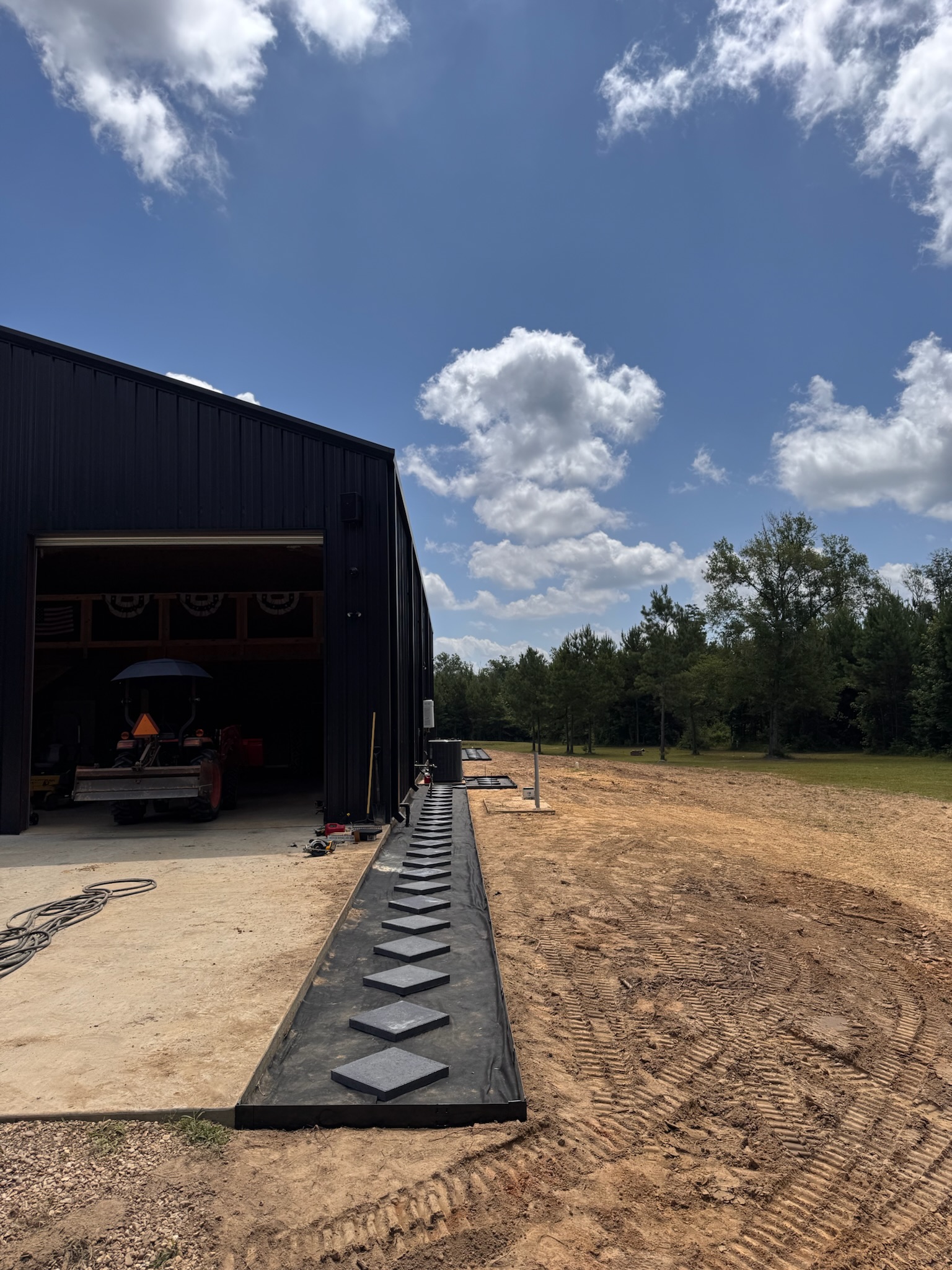 Graded dirt, machine tracks, and a paver path under construction beside a metal shop